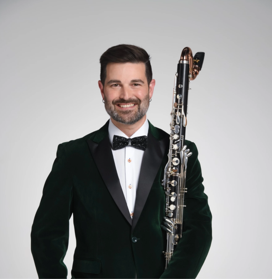 Un hombre de cabello oscuro muy corto, barba y bigote, vestido con un esmoquin negro, sostiene un clarinete bajo en su brazo izquierdo.