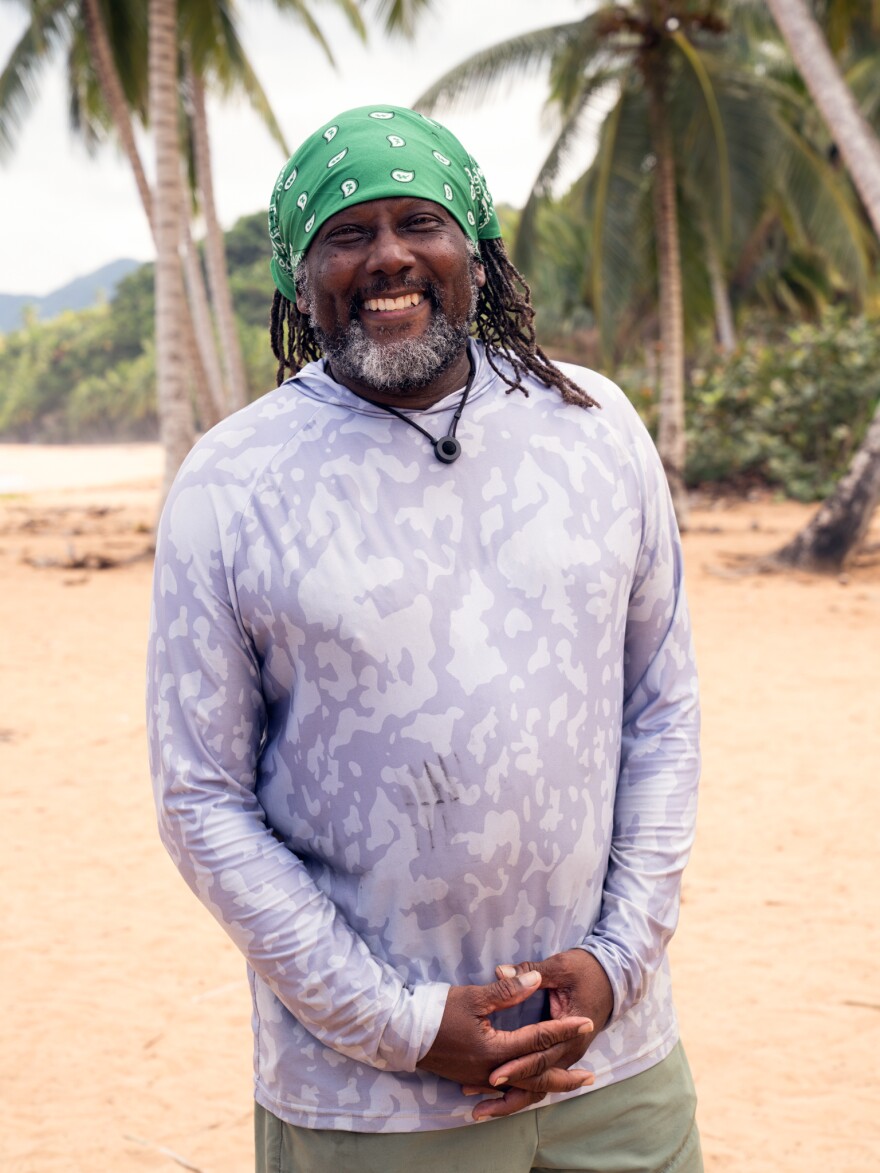 African American man with hands clasped in front of him wearing long sleeve shirt on the beach smiling for a headshot 