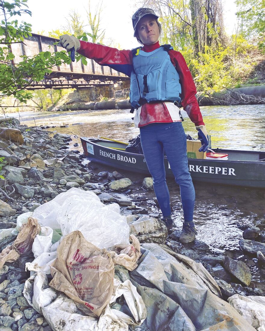 Anna Alsobrook, MountainTrue's Watershed Science and Policy Manager, stands next to a pile of plastic bags that she fished out of the French Broad River. She wears a hat, a red jacket, a blue life jacket, and jeans. A river and bridge is behind her. She is giving a thumbs down to the pile of plastic bags at her feet.