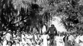 An old black and white photograph shows African Americans gathered around a stage. One man in a suit and hat walks across the stage.