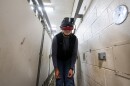 A person wearing a specialized virtual reality headset bends forward while standing in a narrow, indoor corridor with white walls.