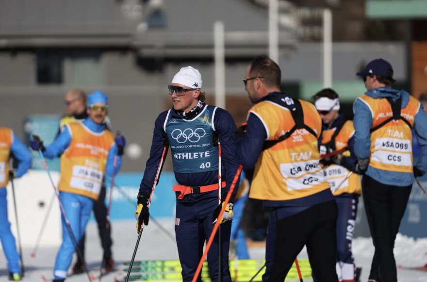 An Olympic cross-country skier wearing sunglasses and spandex stands on alongside team staff.