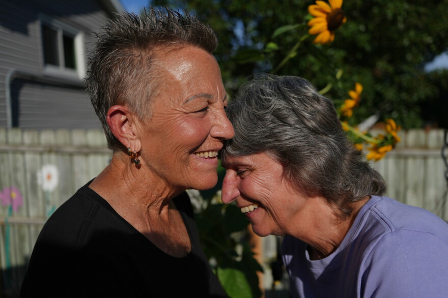2 older white women laugh in the backyard of a home  , the one on the left has short cropped grayish hair in a black top.... the one on the right has darker hair and a light blue top 