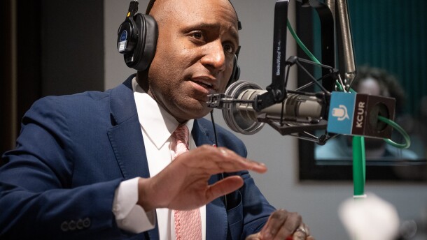 A man in a blue suit jacket with a pink tie gestures with his right hand. He is seated and talking at a microphone inside a radio studio.