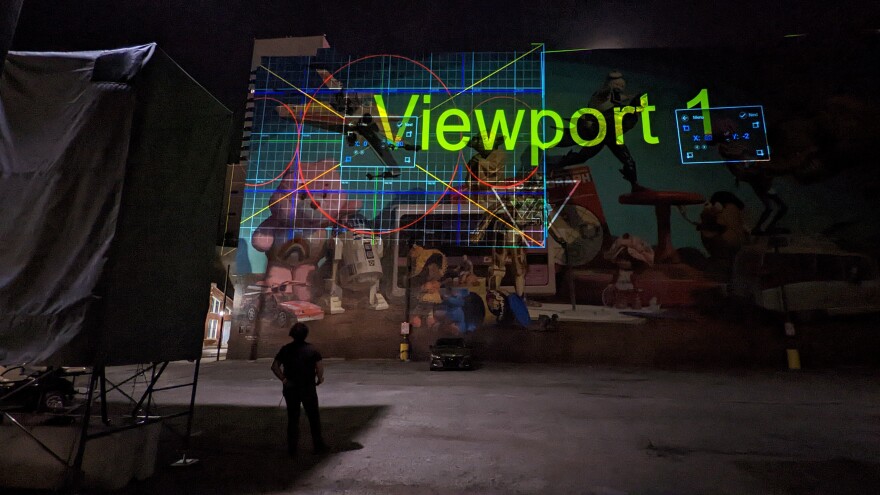 A man stands before the Toy Heritage mural at night. The mural is partially lit with computer graphics displayed on it.