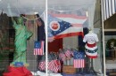 A storefront has a mini Statue of Liberty and many American flags in the window, including several flags that say "250" or '76. The largest flag is an Ohio flag with "250" in the center of the circle. A mannequin wears an America 250 shirt and a skirt made out of red, white, and blue bunting. 