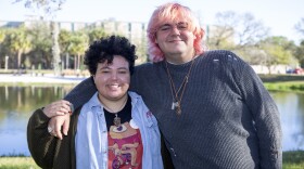 A young afro latino trans man  with short, curly dark hair and wearing a blue button up shirt stands next to a white trans woman with pink hair and a great sweater. They're both smiling.