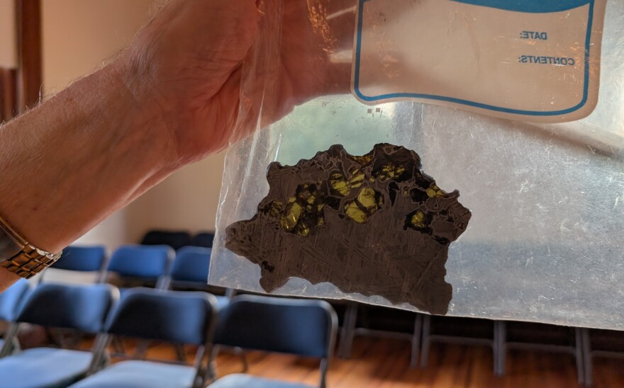 A slice of silvery material in a plastic bag is held against a window. Light comes through part of the space rock.