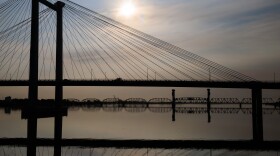 A white cable bridge is silhouetted by the sun. The sky is a pale blue. A river is under the bridge.