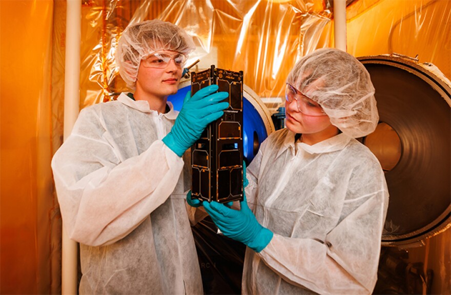 Benjamin Kurtz (left) and Gretta Thompson in a thermal vacuum chamber room, holding a satellite. SAL-E was designed and created by a team of students at the Cal Poly CubeSat Laboratory.