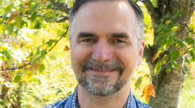A man with brown hair and a short beard wears a plaid blue button down shirt and crosses his arms. He looks into the camera and smiles. There are trees behind him. 