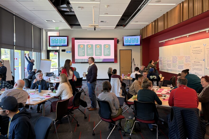 People sit around tables and stand in a room talking.