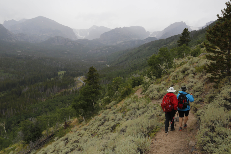 Two hikers walking along a dirt path that's slowly winding down the side of a ridge. There are lots of trees and some mountain peaks in the background.