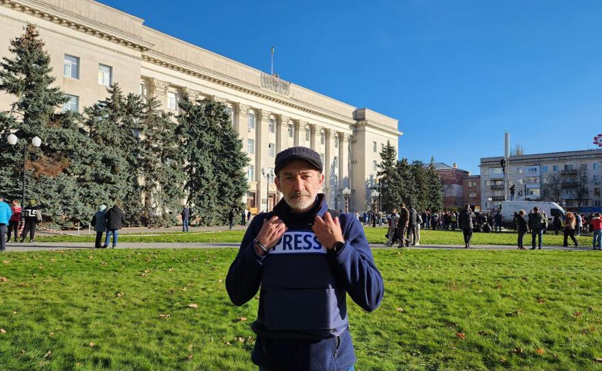 A man wearing a hat and bulletproof vest stands outside.