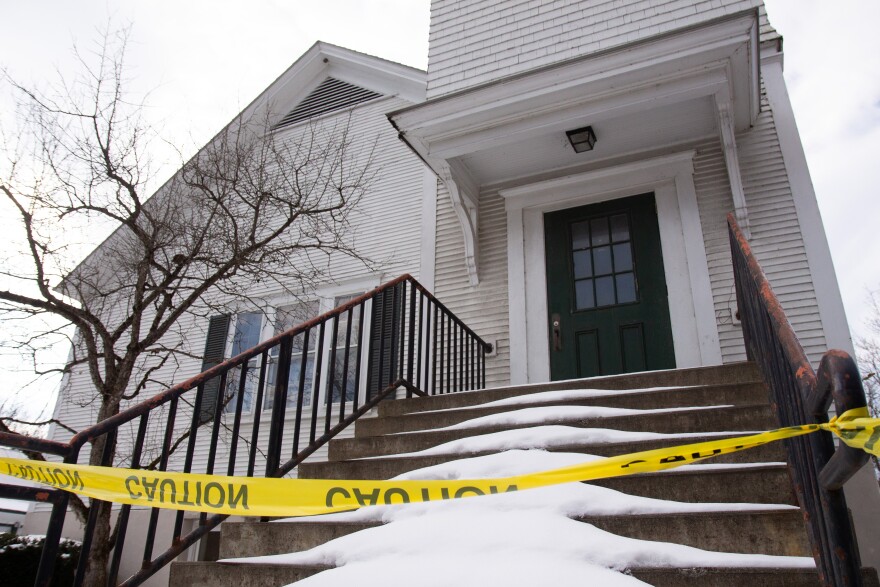 A photo of a white building with a green door and shutters, with cement steps leading up to the door. Yellow caution tape is strung across the front of the steps, which have snow on them.