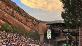 Music fans enjoy a sunset concert at Red Rocks Amphitheater. On the left side is large, tilted rock formation that is a rust-red color with green vegetation. Thousands of fans are seated on a downward slope facing the stage. The band performs on the stage in the lower right corner. There is a big green tree, the sky is blue and sun is setting behind a big cloud.