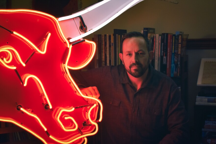 A man sits next to a neon sign in the shape of a bull, lined in glowing red and white neon tubes. 