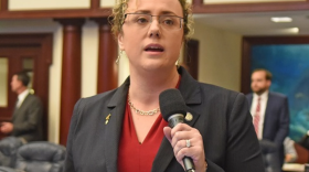 Una mujer con cabello rubio rizado y gafas habla por un micrófono.