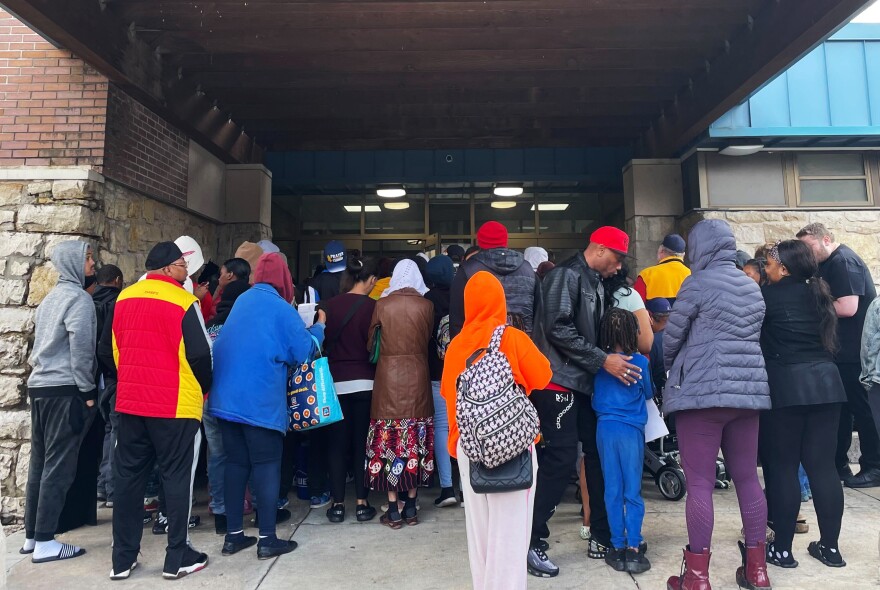 A large crowd of people wearing heavy, cold-weather clothing huddle beneath a canopy outside a building.