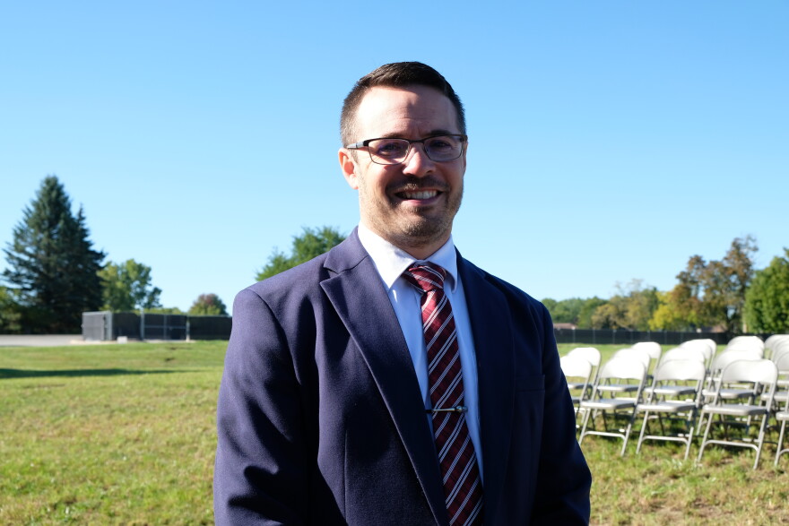 A man in a navy suit and glasses smiles for the camera. There are rows of metal folding chairs on the lawn behind him and black tarp over a chain link fence at the back of the lot. He's standing on the ground where a chapel in the Church of Jesus Christ of Latter Day Saints stood and will be rebuilt again in the coming months.