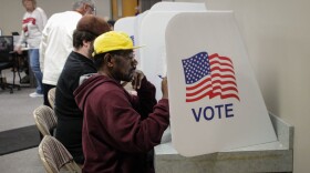 A man in a burgundy jacket and yellow hat sits in a voting booth.