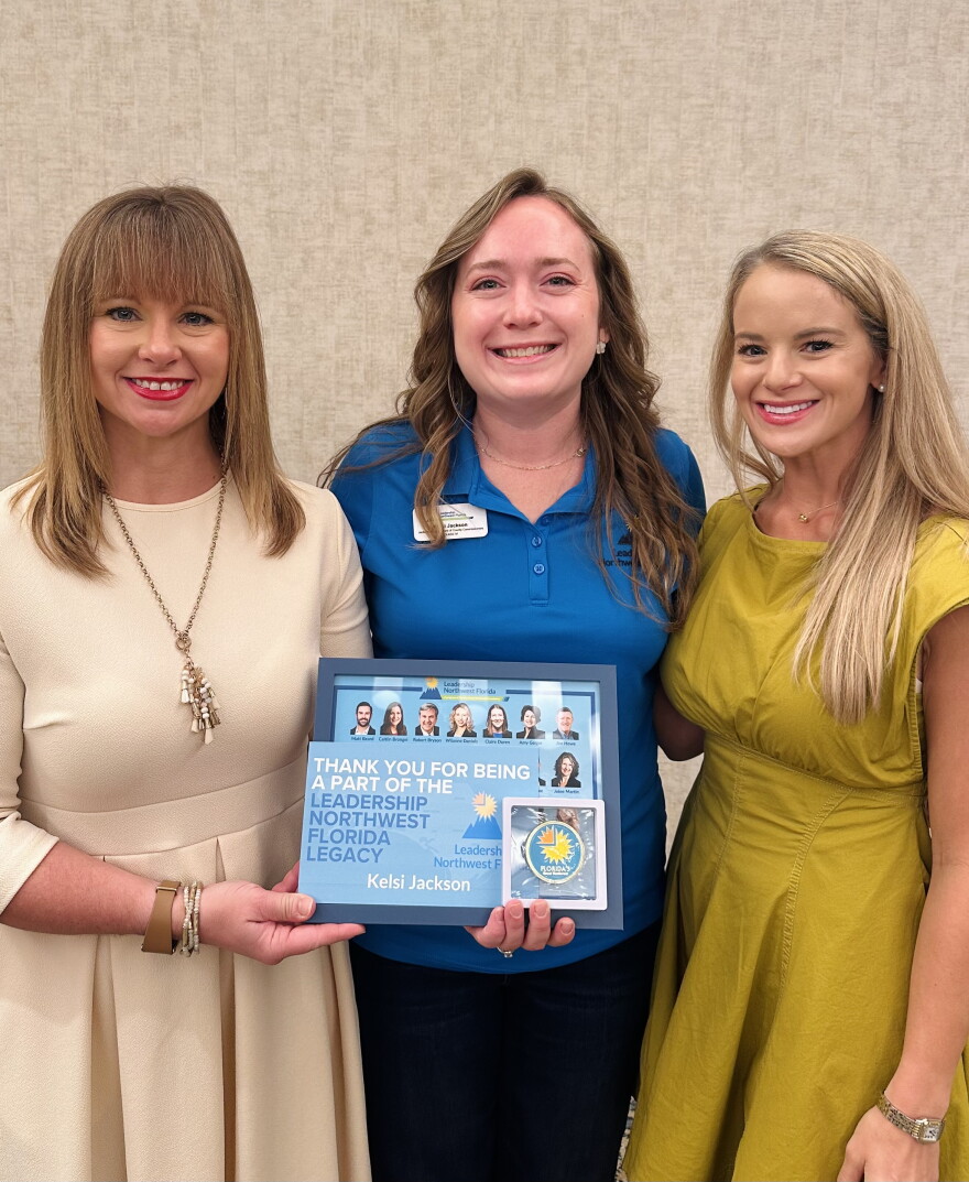 Three women in business casual attire pose with a plaque highlighting Leadership Northwest Florida Legacy for Kelsi Jackson in the middle.