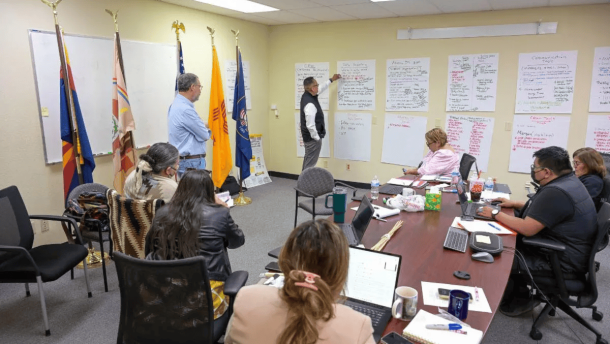 People stand and sit in a room facing a wall with worksheets and posters on it. They are having a work meeting. Flags are hanging on poles in the room.