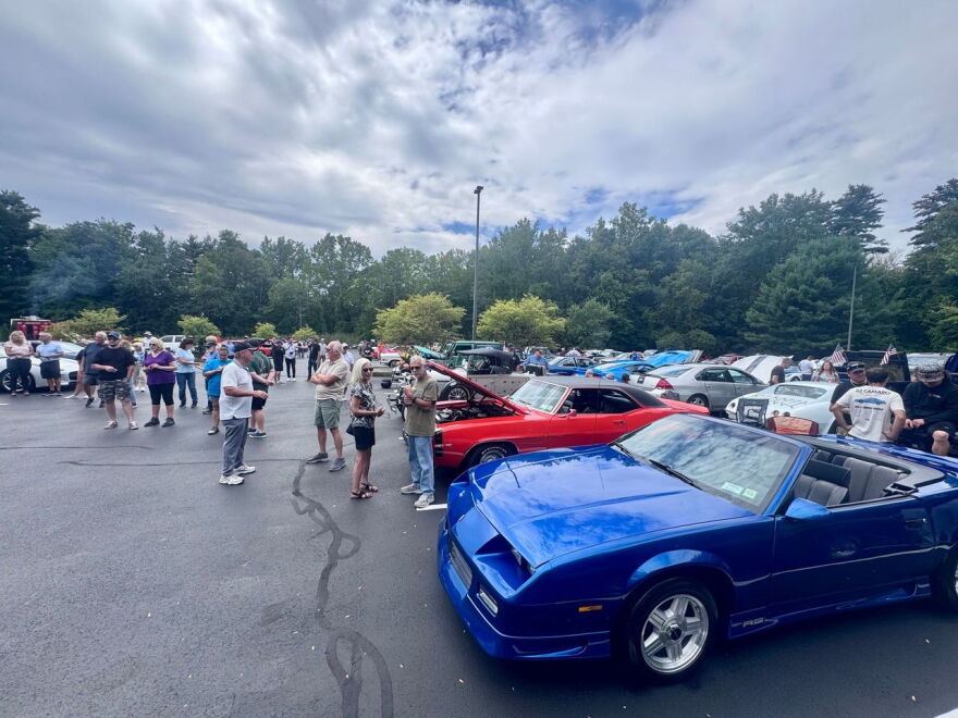 There's rows of muscle cars in a parking lot, which a bunch of people standing around talking. The sky is mostly cloudy, and trees line the parking lot. 