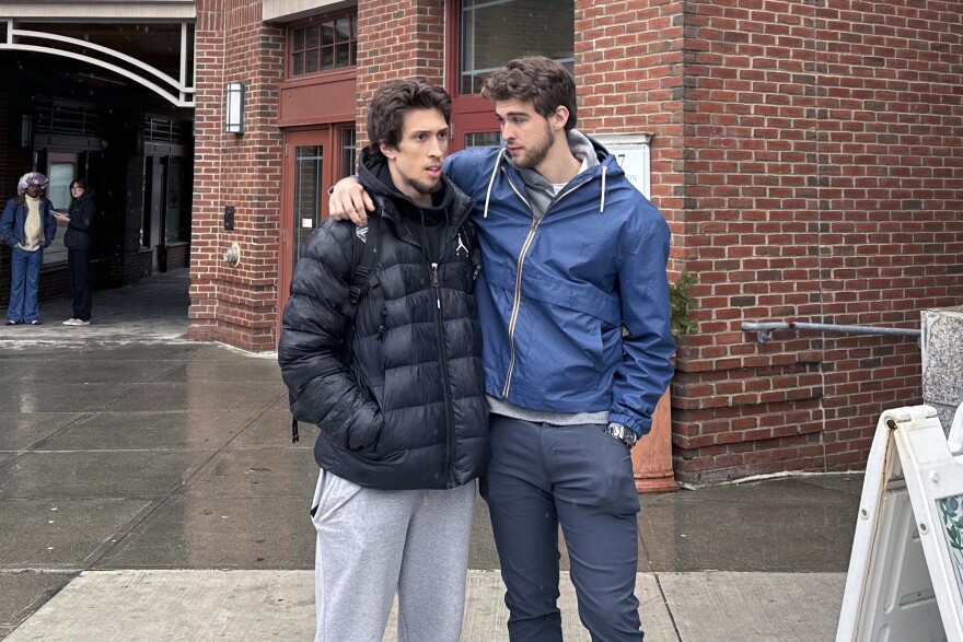 Two young men stand in front of a brick building. One man's arm is draped over the other's shoulders