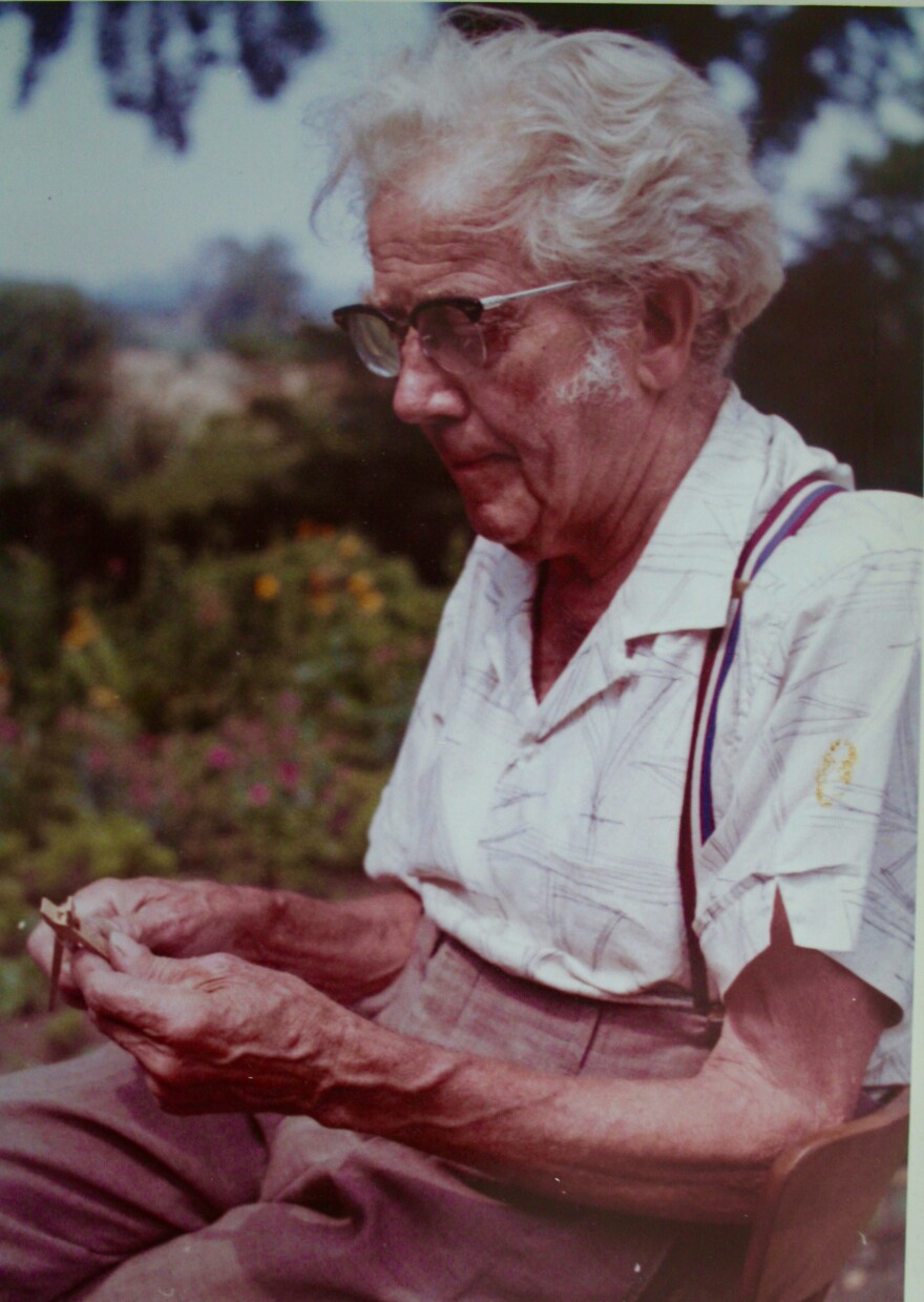 Old man with lots of white hair holds a pair of wooden pliers he's made.