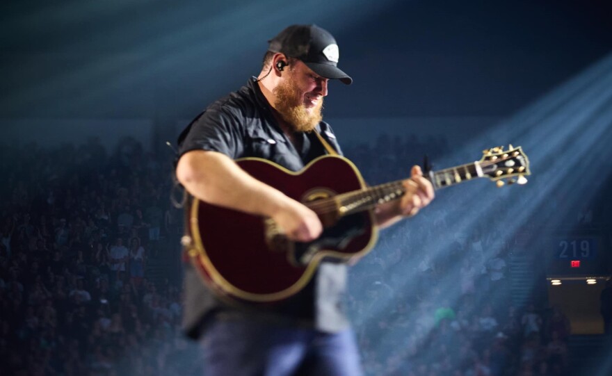 A man with a beard wearing a dark baseball cap and black short-sleeved shirt plays acoustic guitar while walking across the stage during a concert at a large arena.