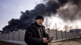 A man holds a mobile phone while smoke rises after Russian attacks hit a fuel storage facility in the city of Kalynivka.