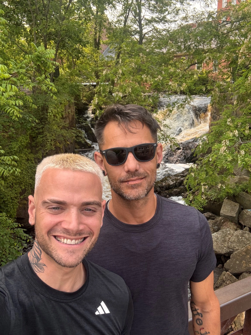 Un hombre rubio y otro castaño se toman una selfie junto a la vegetación y una cascada.