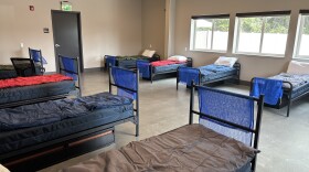 Eight single beds are lined up in a room with a concrete floor and windows. 