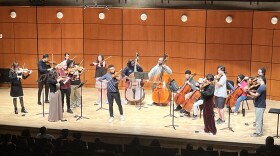 Musicians rehearse onstage in a concert hall, with violinists standing at music stands.