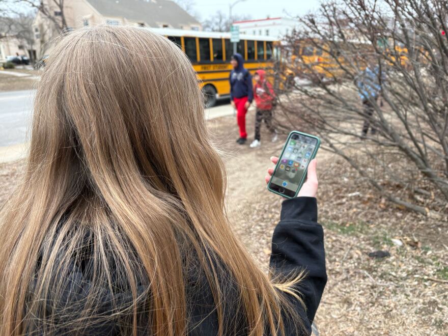 A girl uses a phone with school buses in the background. 