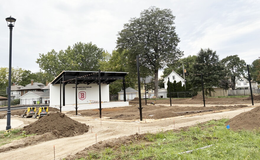 A small outdoor stage sits in a park under construction, with dirt piles and pathways being formed. Several houses and trees are visible in the background.