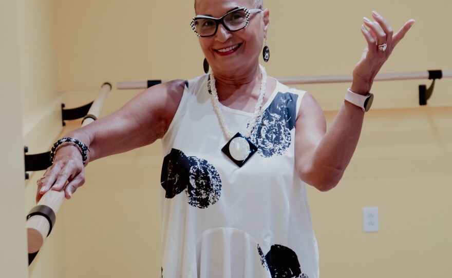 A woman with her grey hair pulled into a ponytail, wearing glasses, bracelets, a watch, and a white dress with black flowers. She is posing in a dance studio. 