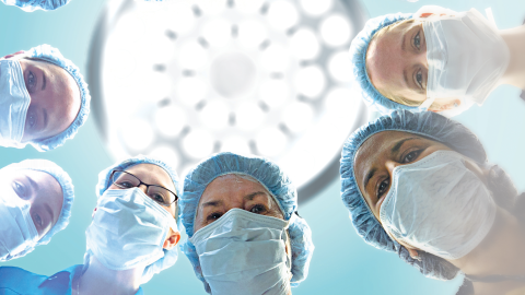 1001 Cuts - Six women in in scrubs and face masks look down at the camera in an operating room with a light above them.