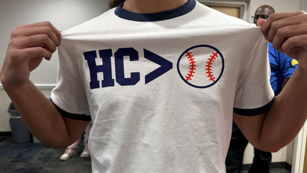A man grabs the sides of his white shirt to showcase the text that reads "HC > Baseball," but the baseball is an icon, and not words.