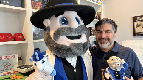 Neal Hoffman stands with the Moshe the Mensch mascot in his home office. He holds a Mensch on a Bench in his hand.