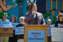 A man stands at a podium and holds a microphone. People behind him hold signs that say "People not politicians."
