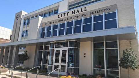 Facade of Garland City Hall. Building is tan and brown with large windows. Yellow flowers and trees ar in front of building. 