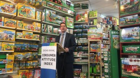 Man stands at podium with sign “Zions Bank Consumer Attitude Index.” Toys line the shelves behind him.