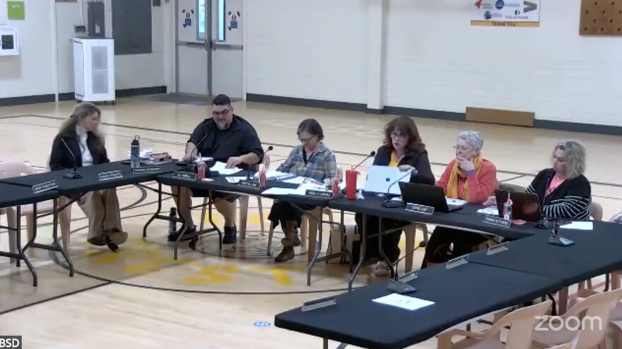 Black folding tables are set up in a school gymnasium. Six people are seated at the tables, with computers, papers, and microphones in front of them. 