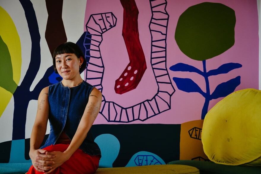 a woman sits with her hands folded on her knees with a tight smile to the camera