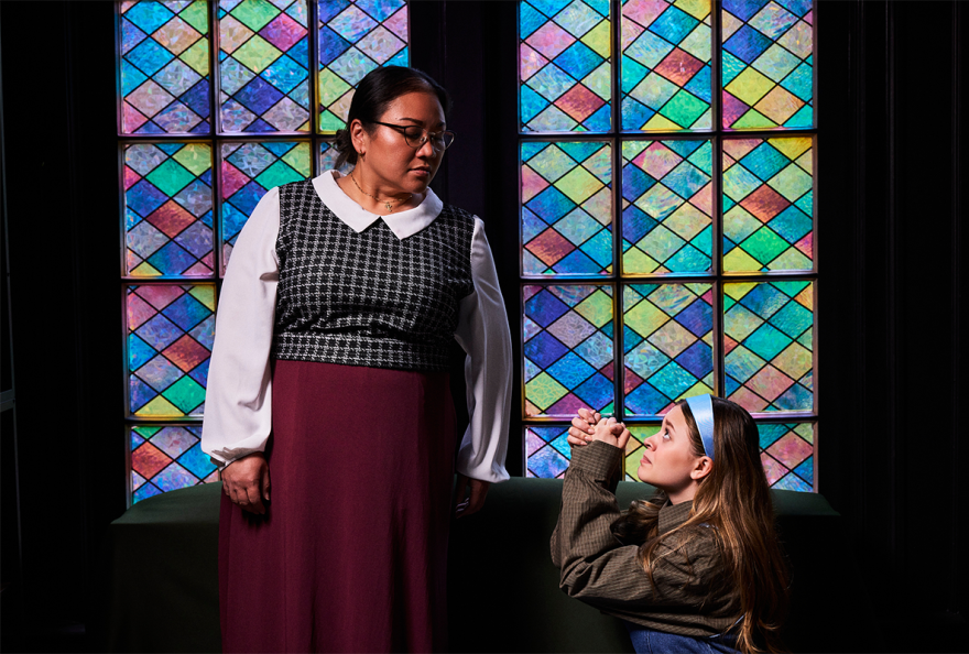 A young woman in a light blue headband kneels with praying hands in front of a woman who looks down at her. They are posed in front of a stained glass window.