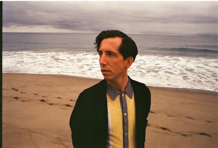 Pokey LaFarge looks off camera into the distance on a beach with the ocean behind him.