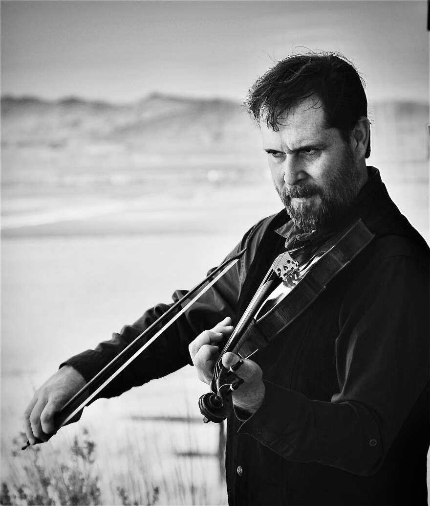 A man with a beard and a serious expression plays the violin in a black and white image.
