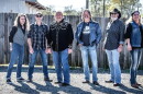 Group of older white men wearing jeans and flannel or button-down shirts standing outside in front of a fence 